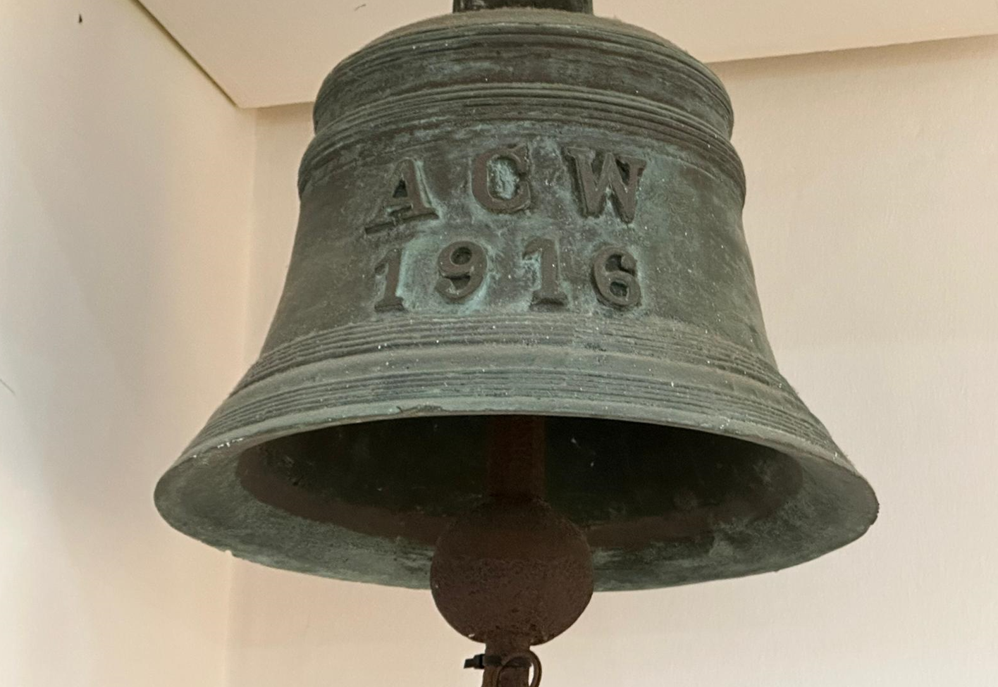 A large green patinated bronze bell inscribed with "ACW 1916" hanging against a white wall.